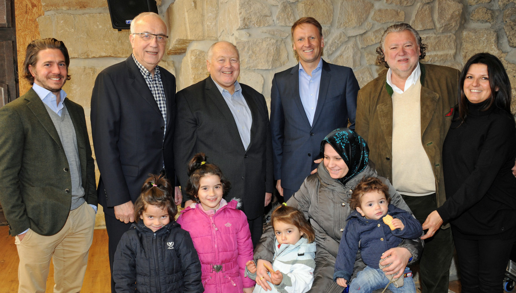 Eine Gruppe von sieben Erwachsenen und vier Kindern posiert für ein Gruppenfoto in einem Raum mit Steinwänden. Die Erwachsenen sind festlich gekleidet, die Kinder tragen Alltagskleidung. Die Atmosphäre wirkt freundlich und gemeinschaftlich.