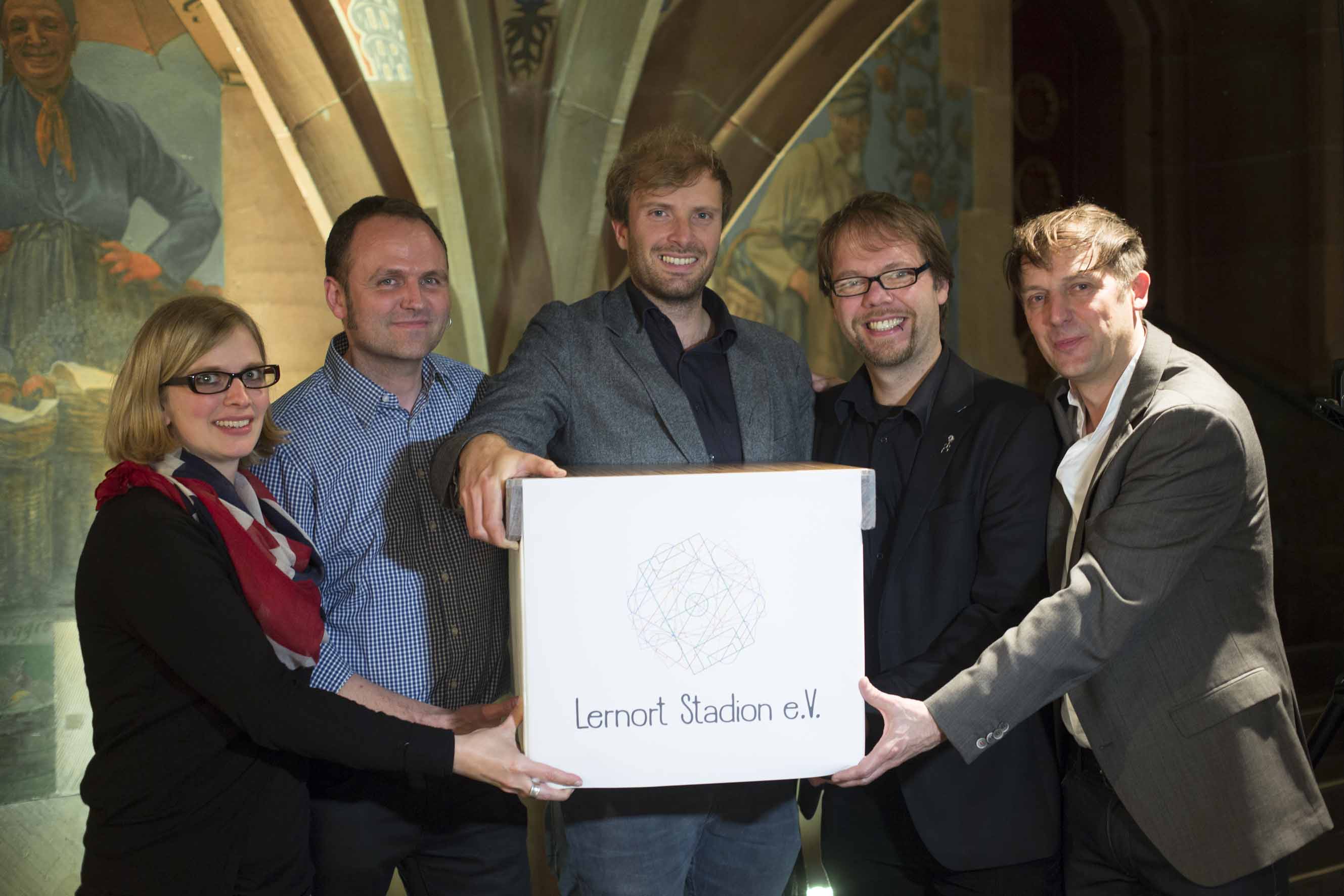 Fünf Personen stehen lächelnd zusammen und halten eine weiße Box mit dem Aufdruck "Lernort Stadion e.V.". Im Hintergrund ist ein historischer Raum mit Gewölbe und Gemälden zu sehen.