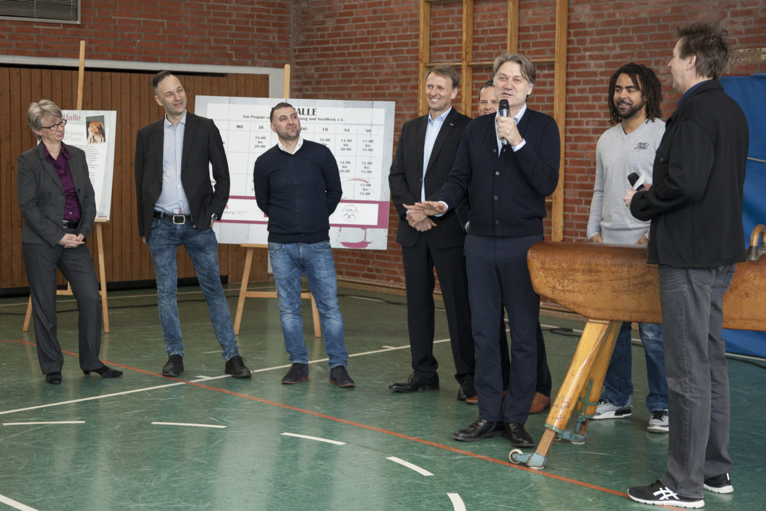 Eine Gruppe von Menschen steht in einer Turnhalle zusammen. Im Hintergrund sind Plakate zu sehen. Ein Mann hält ein Mikrofon und spricht. Die anderen Zuhörer stehen oder sitzen und schauen ihm zu.