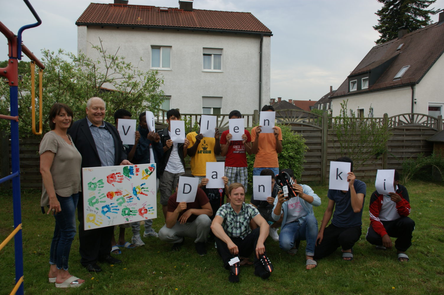 Eine Gruppe von Jugendlichen und Erwachsenen steht auf einer grünen Wiese vor einem Zaun und Häusern. Einige halten Buchstaben vor ihre Gesichter, die zusammen den Begriff "Denken" ergeben. Ein junges Mädchen hält ein bemalten Karton hoch.