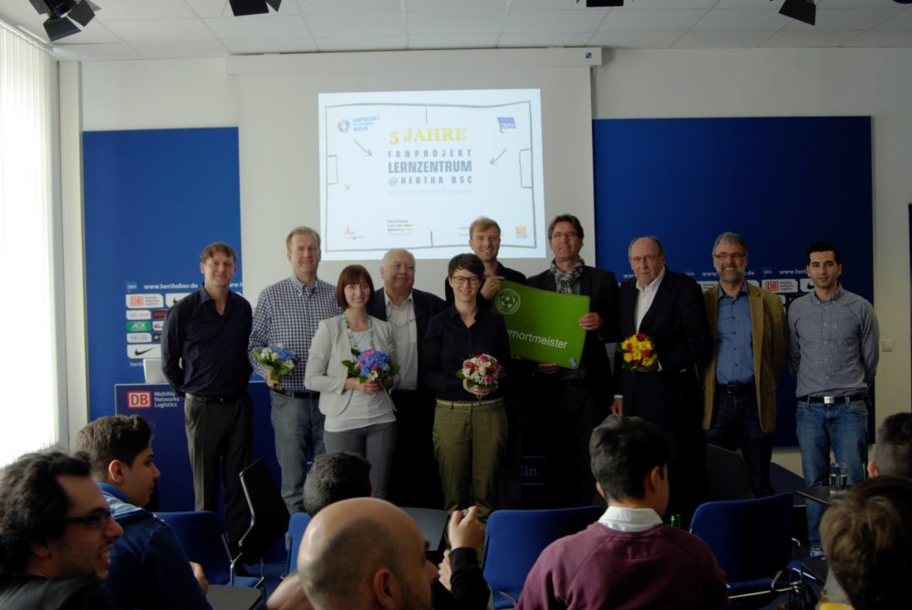 Gruppenfoto von Personen bei der Übergabe einer Plakette oder eines Schildes, vermutlich im Rahmen der Eröffnung oder Erweiterung eines Lernzentrums in Berlin. Im Hintergrund eine Projektionsfläche mit dem Hinweis "10 Jahre Lernzentrum Berlin".