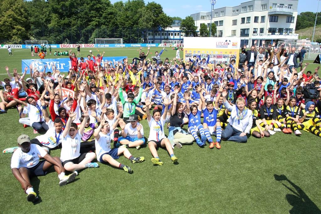 Eine große Gruppe von Jugendlichen und Betreuern steht und sitzt auf einem Fußballplatz und lächelt in die Kamera. Viele tragen T-Shirts in verschiedenen Farben und einige tragen Fußballtrikots. Im Hintergrund ist ein Fußballstadion zu sehen.