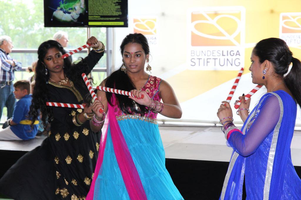 Drei junge Frauen in bunten traditionellen Kleidern tanzen mit bunten Bändern, im Hintergrund ein Banner mit dem Logo der DFL Stiftung.