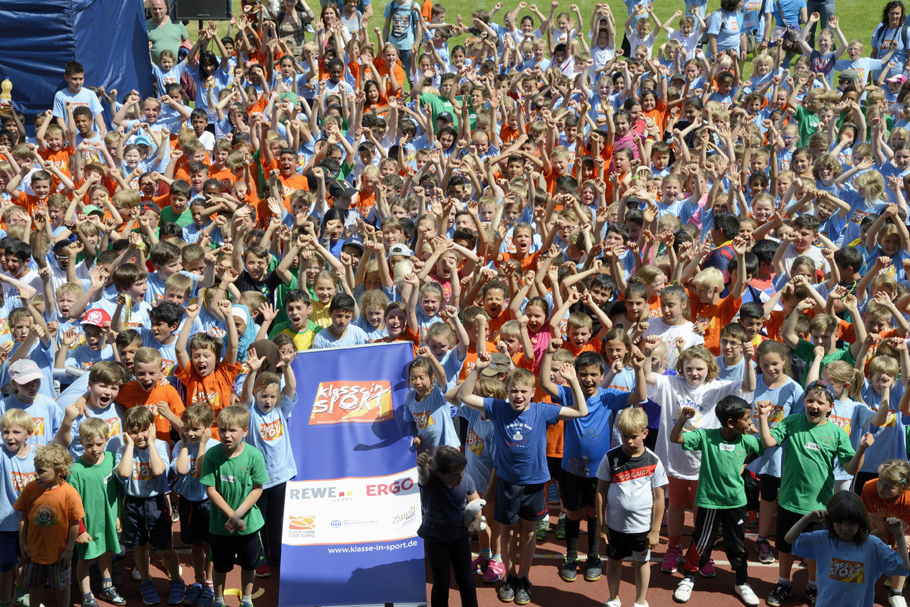 Jubelnde Kindergruppe beim "Klasse im Sport"-Turnier der DFL Stiftung. Im Hintergrund ein Banner mit den Logos von REWE und ERGO.