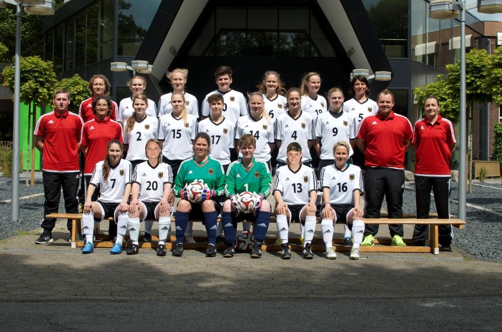 Frauenfußballmannschaft in weißen Trikots und roten Trainingsjacken posiert für ein Mannschaftsfoto vor einem modernen Gebäude. Einige Spielerinnen sitzen auf einer Bank, andere stehen dahinter. Im Vordergrund hält eine Spielerin einen Fußball.