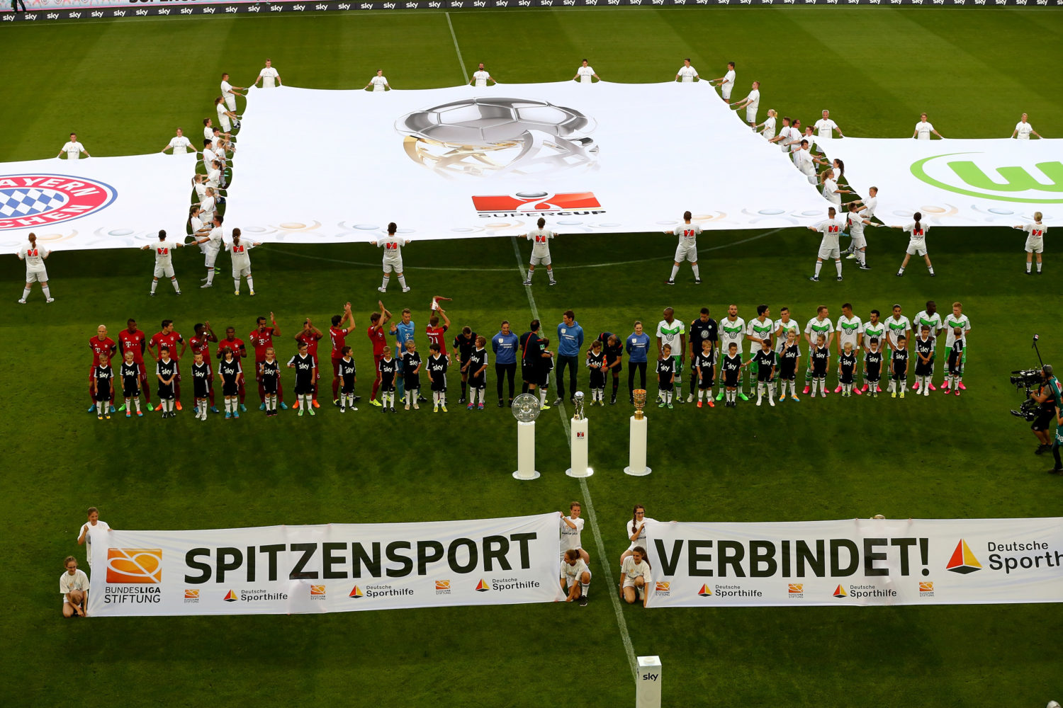 Spieler beider Mannschaften formen vor dem Spiel einen gemeinsamen Gang, während eine große Banner mit dem Aufdruck "Spitzensport verbindet!" präsentiert wird. Im Hintergrund ist das Spielfeld und die Stadiontribüne zu sehen.