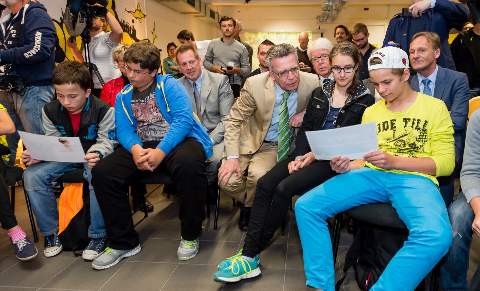 Eine Gruppe von Jugendlichen und Erwachsenen sitzt auf schwarzen Stühlen in einem Raum. Einige der Jugendlichen halten Blätter Papier in der Hand und wirken aufmerksam. Im Hintergrund sind weitere Personen zu sehen.