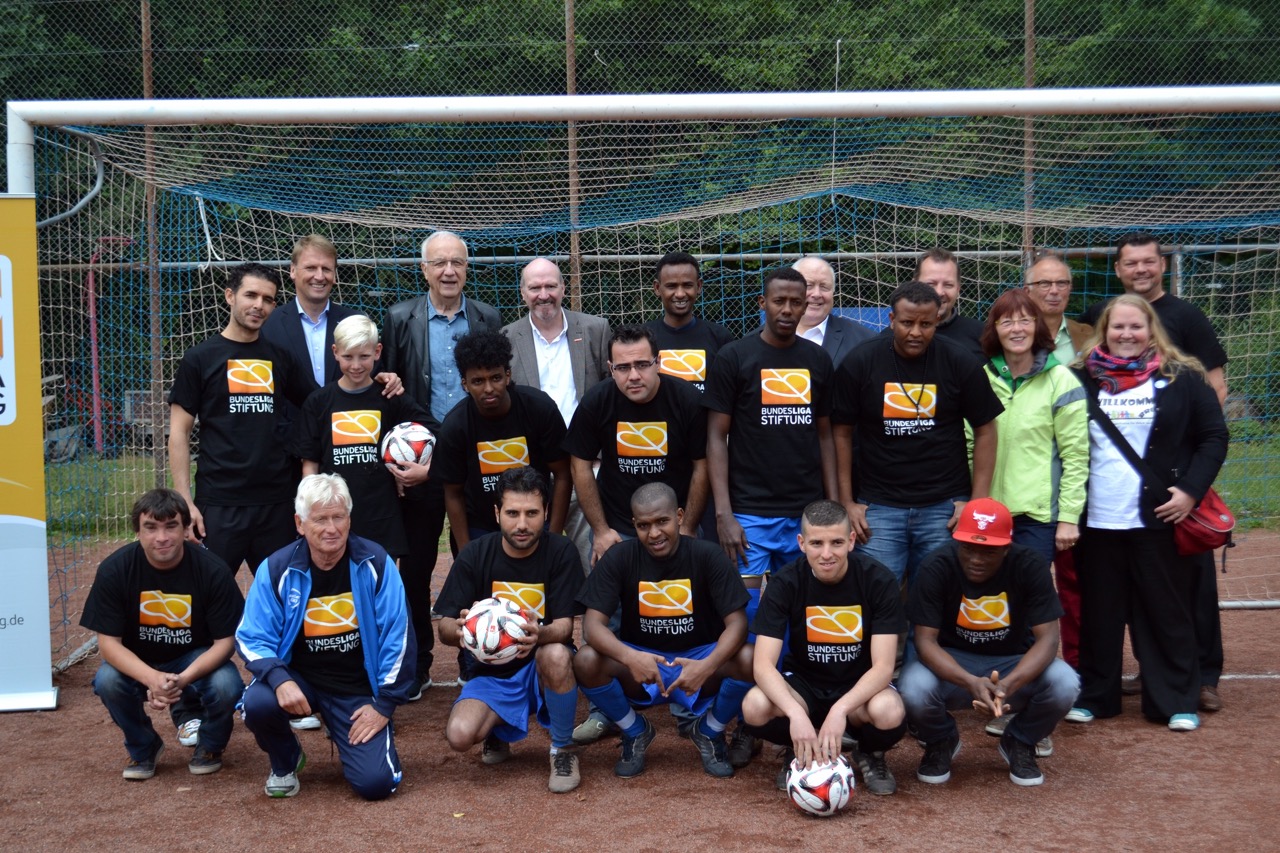Eine Gruppe von Menschen steht auf einem Fußballplatz vor einem Tor. Die meisten tragen T-Shirts mit dem Aufdruck "Bundesliga Stiftung" und halten Fußbälle. Es handelt sich vermutlich um Teilnehmer oder Unterstützer eines Projekts der DFL Stiftung.