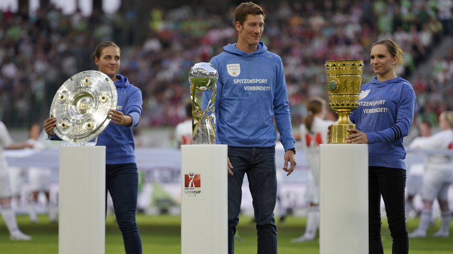 Zwei Frauen und ein Mann in blauen Trainingsanzügen mit dem Aufdruck "SPITZENSPORT VERBINDET!" stehen auf einem Fußballfeld und halten jeweils einen Pokal hoch. Im Hintergrund ist ein volles Stadion zu sehen.
