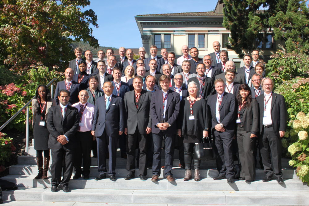 Eine große Gruppe von Personen in formeller Geschäftskleidung posiert auf einer breiten Treppe vor einem hellen Gebäude mit Hecken und Bäumen im Hintergrund. Viele tragen Namensschilder und wirken wie Teilnehmer einer Konferenz oder eines Treffens.