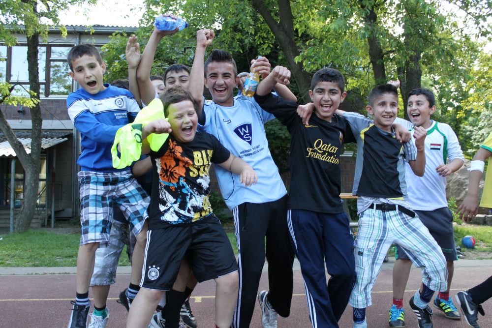 Eine Gruppe jubelnder Jugendlicher steht auf einem roten Sportplatz und freut sich in die Kamera. Einige tragen Fußballtrikots, andere lockere Kleidung. Im Hintergrund ist ein Teil eines Schulgebäudes zu sehen.