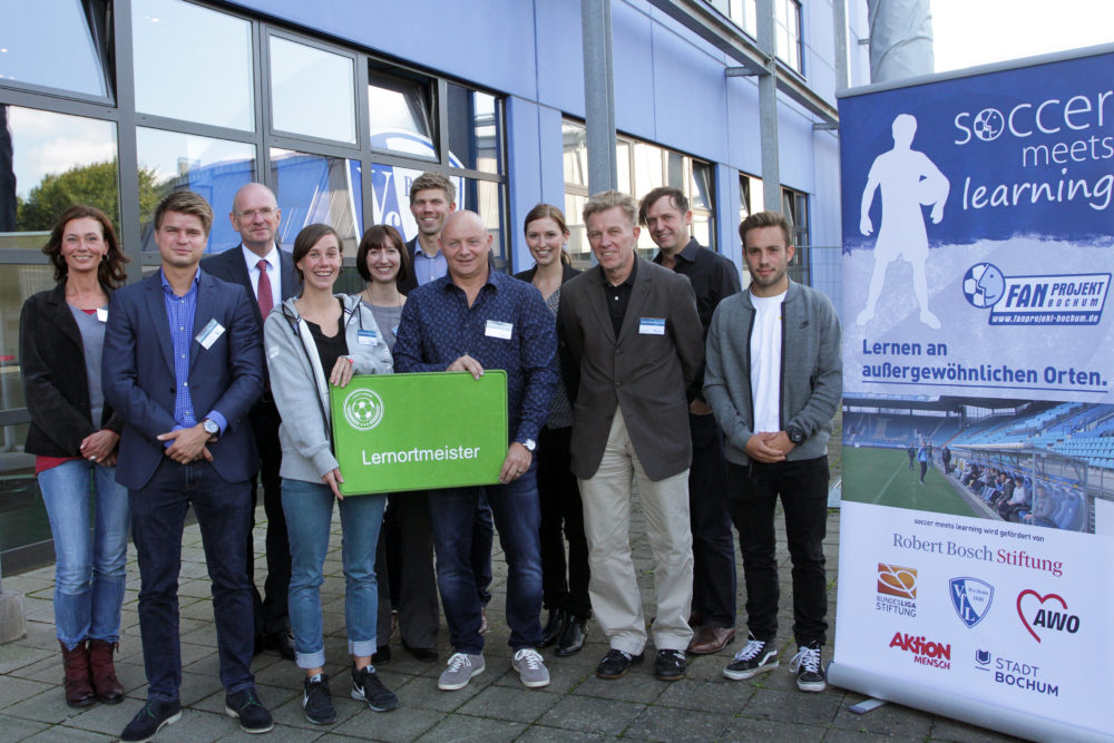 Eine Gruppe von etwa zehn Personen steht lächelnd vor einer gläsernen Tür und einem Banner mit der Aufschrift "Soccer meets Learning". Im Vordergrund hält ein Mann ein Schild mit der Aufschrift "Lernortmeister". Im Hintergrund ist ein Fußballstadion abgebildet. Das Bild zeigt eine Veranstaltung der DFL Stiftung, die Bildung und Fußball verbindet.