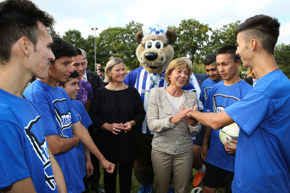Eine Gruppe Jugendlicher in blauen Fußballtrikots steht im Kreis um zwei Frauen und eine Maskottchenfigur. Die Frauen scheinen die Jugendlichen zu begrüßen. Im Hintergrund ist eine Sportanlage zu erkennen.
