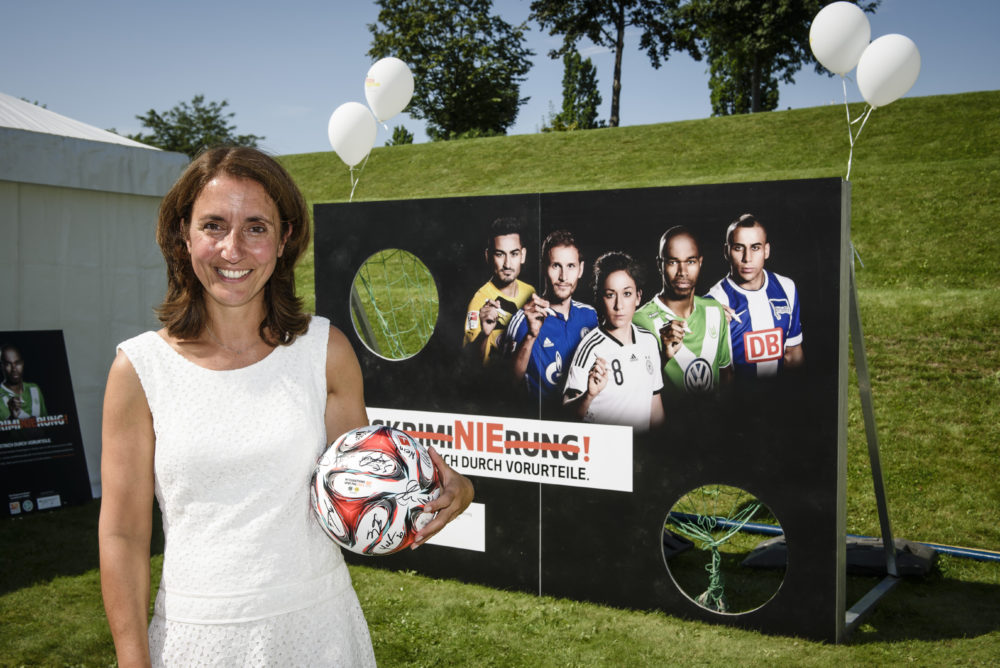 Staatsministerin Aydan Özoguz hält einen Fußball mit Unterschriften vor einem Werbebanner mit Fußballspielern und dem Slogan "Zusammen. Spielend gegen Vorurteile". Im Hintergrund grüne Rasenfläche.