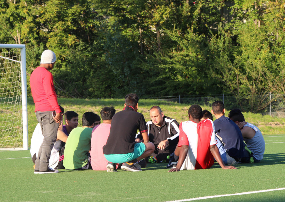 Eine Gruppe von Jugendlichen sitzt auf einem grünen Kunstrasenplatz zusammen und lauscht einem Trainer, der ihnen etwas erklärt. Im Hintergrund steht ein Fußballtor und dichtes Grün.
