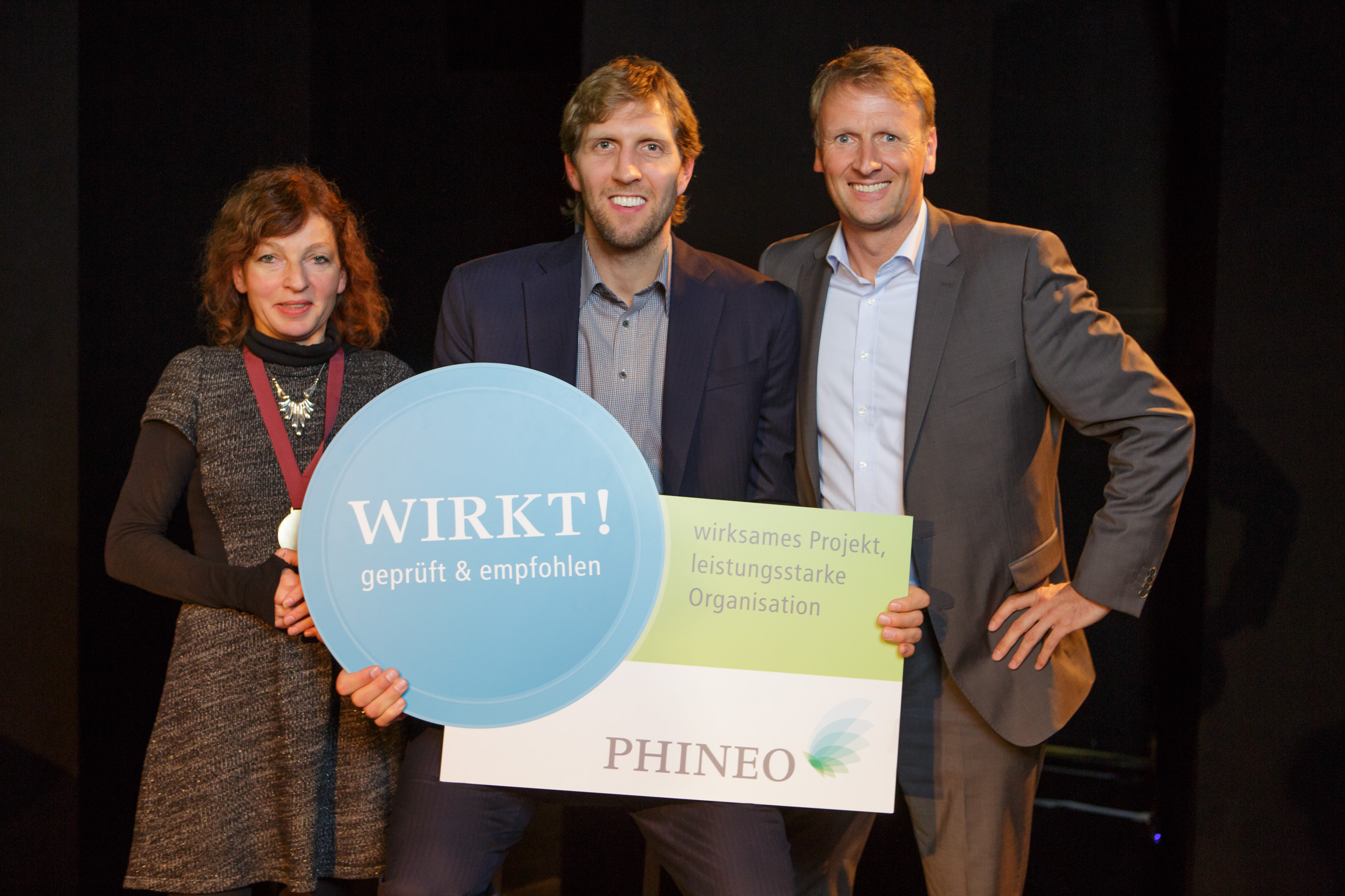 Drei Personen, eine Frau und zwei Männer, halten ein Schild mit dem Aufdruck "WIRKT! geprüft & empfohlen" und dem Logo von PHINEO. Sie lächeln in die Kamera.