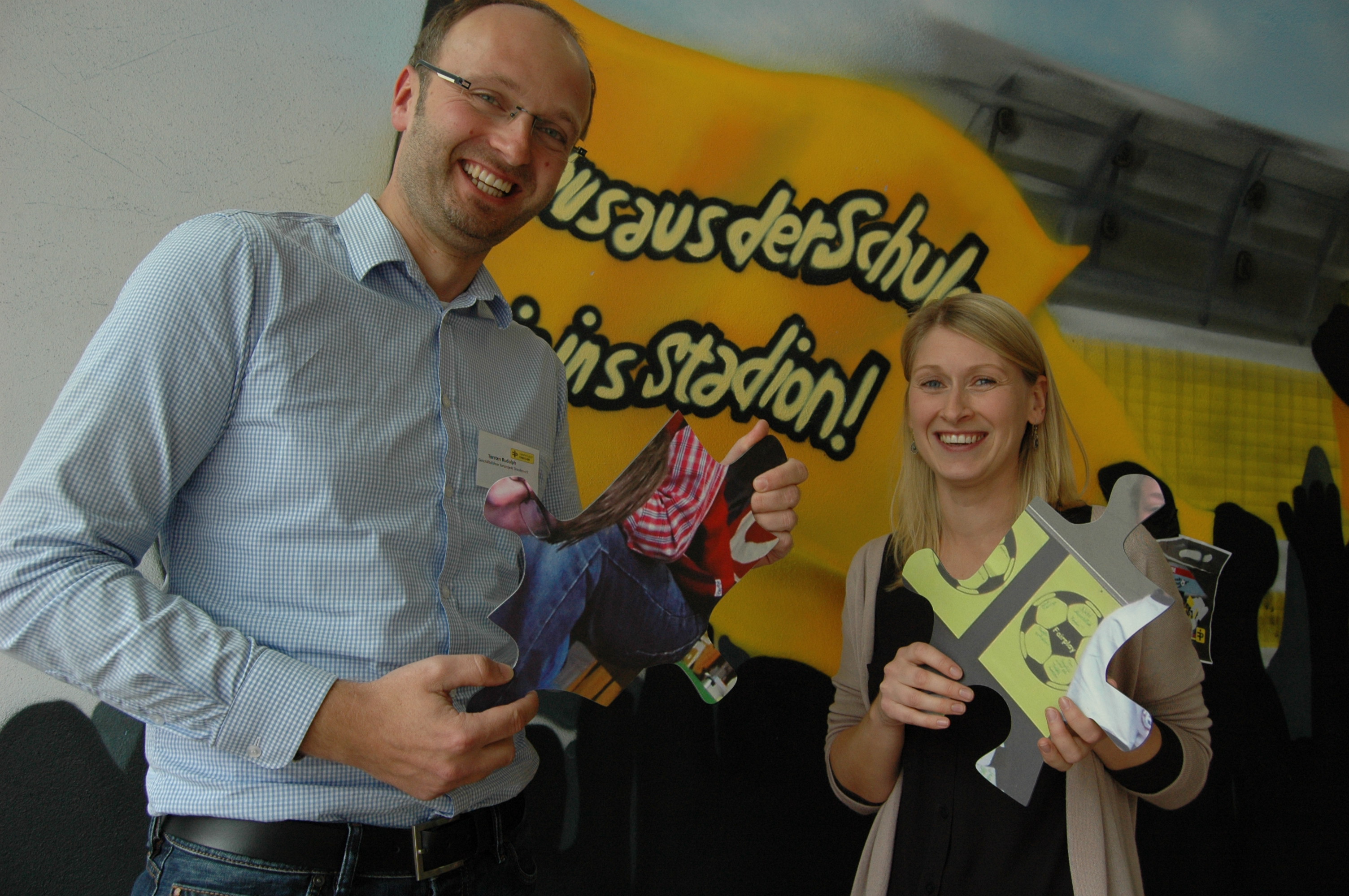 Zwei lächelnde Personen, ein Mann und eine Frau, stehen vor einem Werbebanner mit der Aufschrift "Aus der Schul' ins Stadion!". Die Frau hält zwei Informationsbroschüren in der Hand.