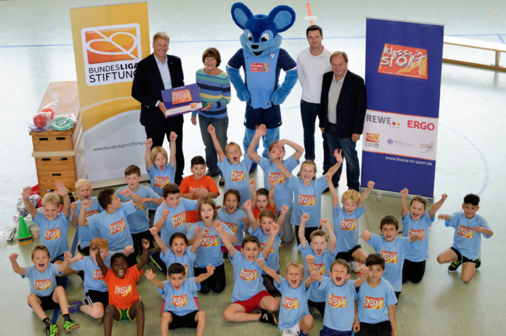 Eine Gruppe von Kindern in blauen T-Shirts mit dem Logo der DFL Stiftung steht in einer Sporthalle und lächelt in die Kamera. Hinter ihnen stehen vier Erwachsene, darunter auch das Maskottchen der DFL Stiftung. Im Hintergrund sind Werbebanner von REWE und ERGO zu sehen.