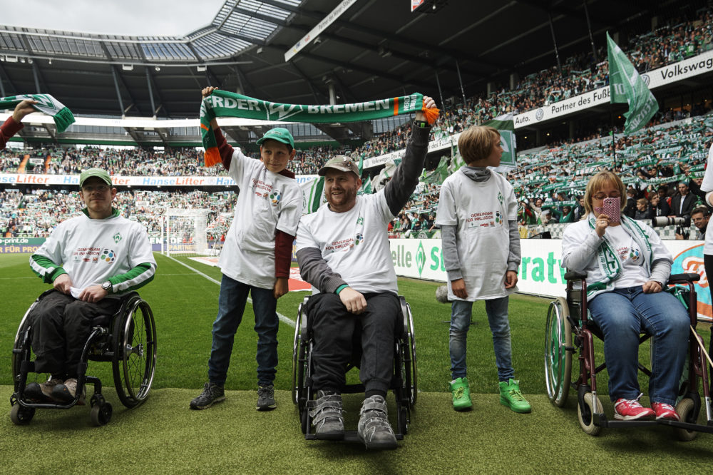 Eine Gruppe von Kindern und Jugendlichen, einige im Rollstuhl, steht auf dem Rasen eines Fußballstadions. Sie tragen weiße T-Shirts und grüne Caps oder Schals der Farben von Werder Bremen und Bayer 04 Leverkusen. Im Hintergrund sind die Zuschauerreihen des Stadions zu sehen. Das Bild zeigt eine inklusive Aktion der DFL Stiftung.