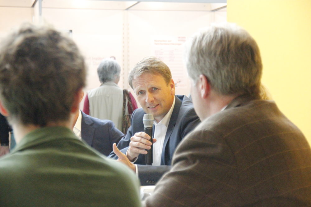 Ein Mann im Anzug hält ein Mikrofon und spricht in einer Gesprächsrunde. Um ihn herum sitzen weitere Personen, die ihm zuwenden.