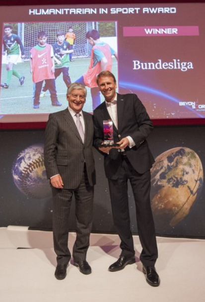 Zwei Männer im Anzug stehen auf einer Bühne. Der Mann links überreicht dem Mann rechts eine gläserne Trophäe. Im Hintergrund ist eine Großbildleinwand zu sehen, die Kinder beim Fußballspielen zeigt und den Text "WINNER Bundesliga" enthält.