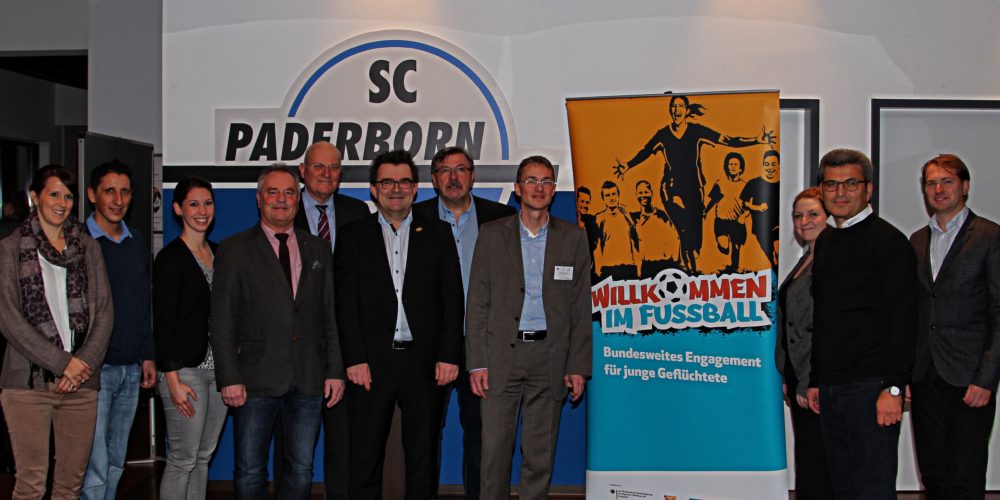 Eine Gruppe von etwa 15 Personen steht vor einem Banner mit dem Aufdruck "WILLKOMMEN IM FUSSBALL – Bundesweites Engagement für junge Flüchtlinge". Das Banner zeigt spielende Kinder verschiedener Hautfarben. Die Personen auf dem Bild sind gemischt nach Geschlecht und Alter und tragen formelle Kleidung. Im Hintergrund ist das Logo von SC Paderborn zu sehen.