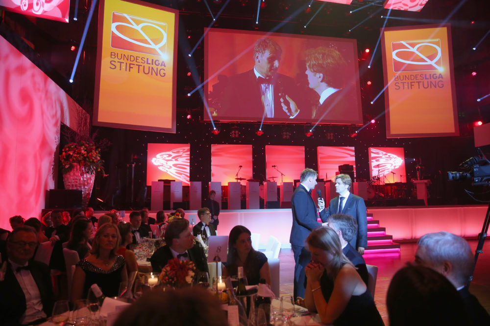 Zwei Männer in dunklen Anzügen stehen auf einer Bühne vor einem Publikum in einem festlichen Saal. Im Hintergrund sind große Bildschirme mit dem Logo der DFL Stiftung zu sehen. Der Raum ist festlich beleuchtet und mit Blumen dekoriert.