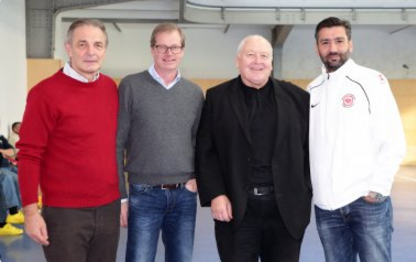 Vier Männer stehen in einer Turnhalle zusammen. Von links: Ein Mann mit rotem Pullover, ein Mann mit grauem Pullover, ein Mann im schwarzen Mantel und ein Mann mit weißer Jacke.