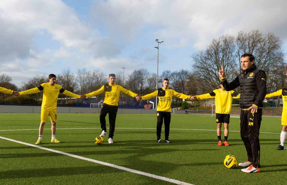 Fußballtrainer gestikuliert während einer Übungseinheit mit Jugendlichen auf einem grünen Kunstrasenfeld. Die Jugendlichen stehen im Kreis und führen Dehnübungen durch.