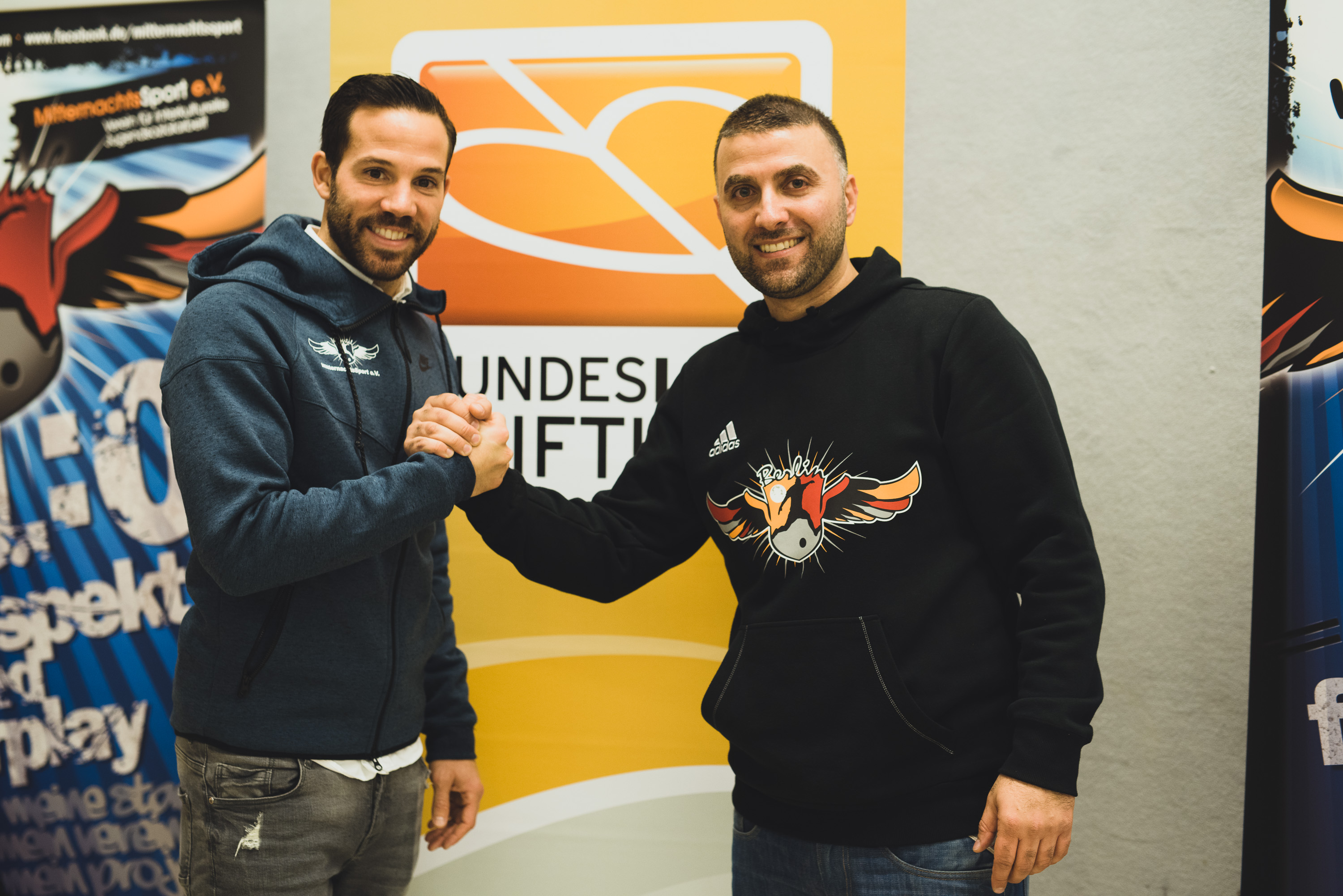 Zwei Männer im Hoodie stehen sich gegenüber und geben sich die Hand. Im Hintergrund ist ein Banner mit der Aufschrift "Mitternachtssport" und das Logo der DFL Stiftung zu sehen.
