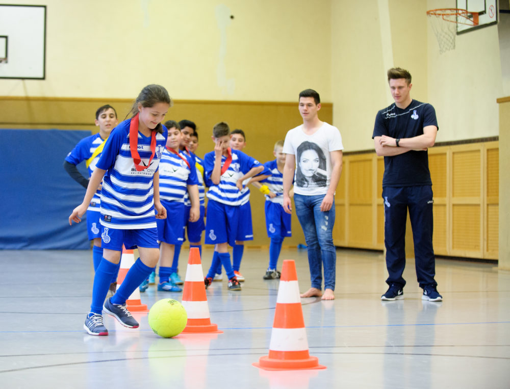 Eine Gruppe von Kindern in Fußballtrikots absolviert einen Dribbelparcours in einer Turnhalle. Ein Junge dribbelt den Ball an Hütchen vorbei, während ein Trainer oder Betreuer im Hintergrund steht und zuschaut.