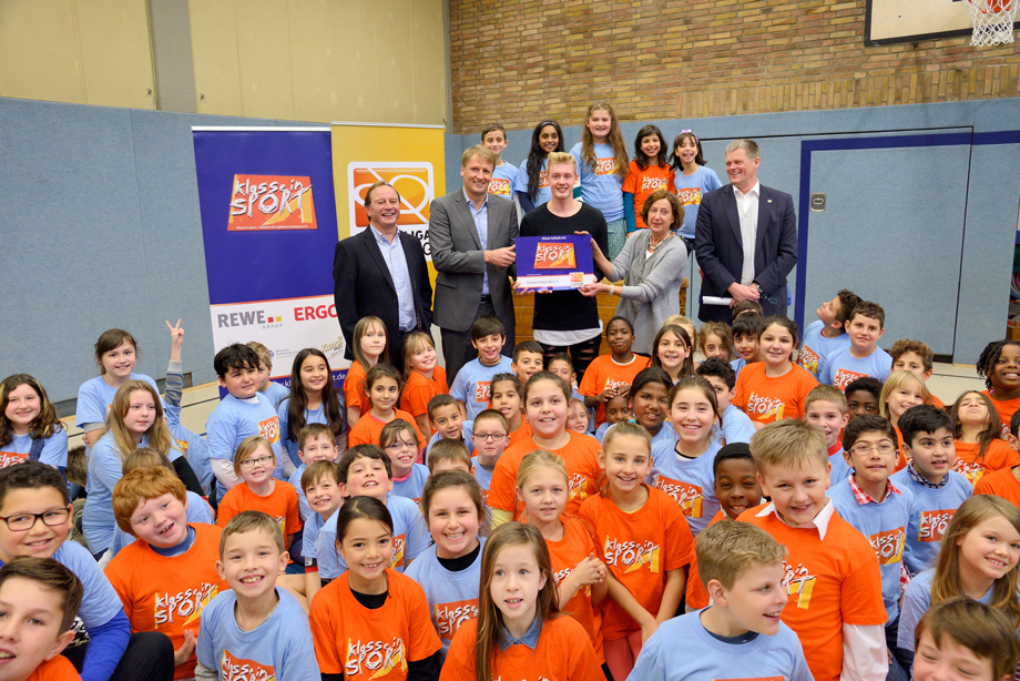 Eine Gruppe von Kindern und Jugendlichen in orangefarbenen und hellblauen T-Shirts posiert mit einem Mann, der eine große Scheckübergabe hält. Im Hintergrund stehen weitere Personen, vermutlich Vertreter der DFL Stiftung und Förderer. Das Bild zeigt eine Veranstaltung im Rahmen des Projekts "KISS - Kleine integrierte Sportgruppen".