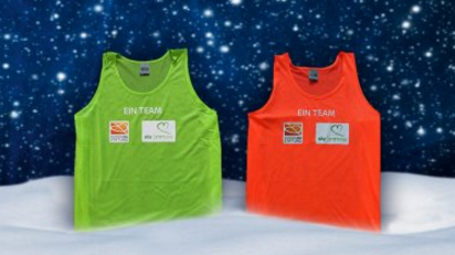 Zwei Sport-Tanktops, eines grün und eines rot, mit dem Aufdruck "EIN TEAM" und dem Logo der DFL Stiftung vor winterlichem Hintergrund.