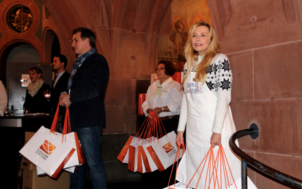Eine Frau in einem weißen Strickkleid und ein Mann im Anzug stehen auf einer Treppe. Beide tragen orangefarbene Tragetaschen mit dem Logo der DFL Stiftung. Im Hintergrund sind weitere Personen und ein festlicher Raum erkennbar.
