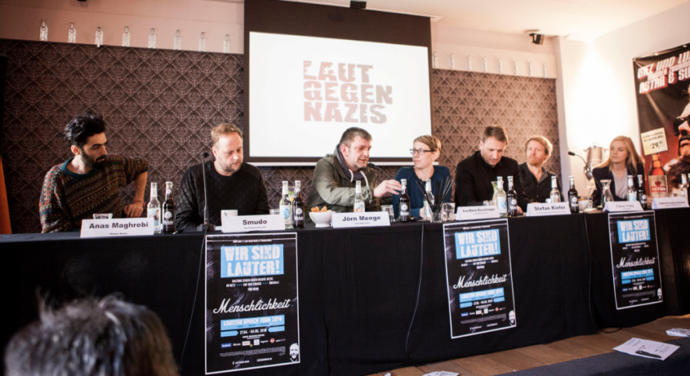 Eine Gruppe von Menschen sitzt an einem Tisch vor einem Banner mit der Aufschrift "GEGEN NAZIS". Auf dem Tisch stehen Schilder mit dem Text "WIR SIND LAUTER! Menschlichkeit". Die Teilnehmer*innen sind Teil einer Diskussionsrunde oder eines Panels.