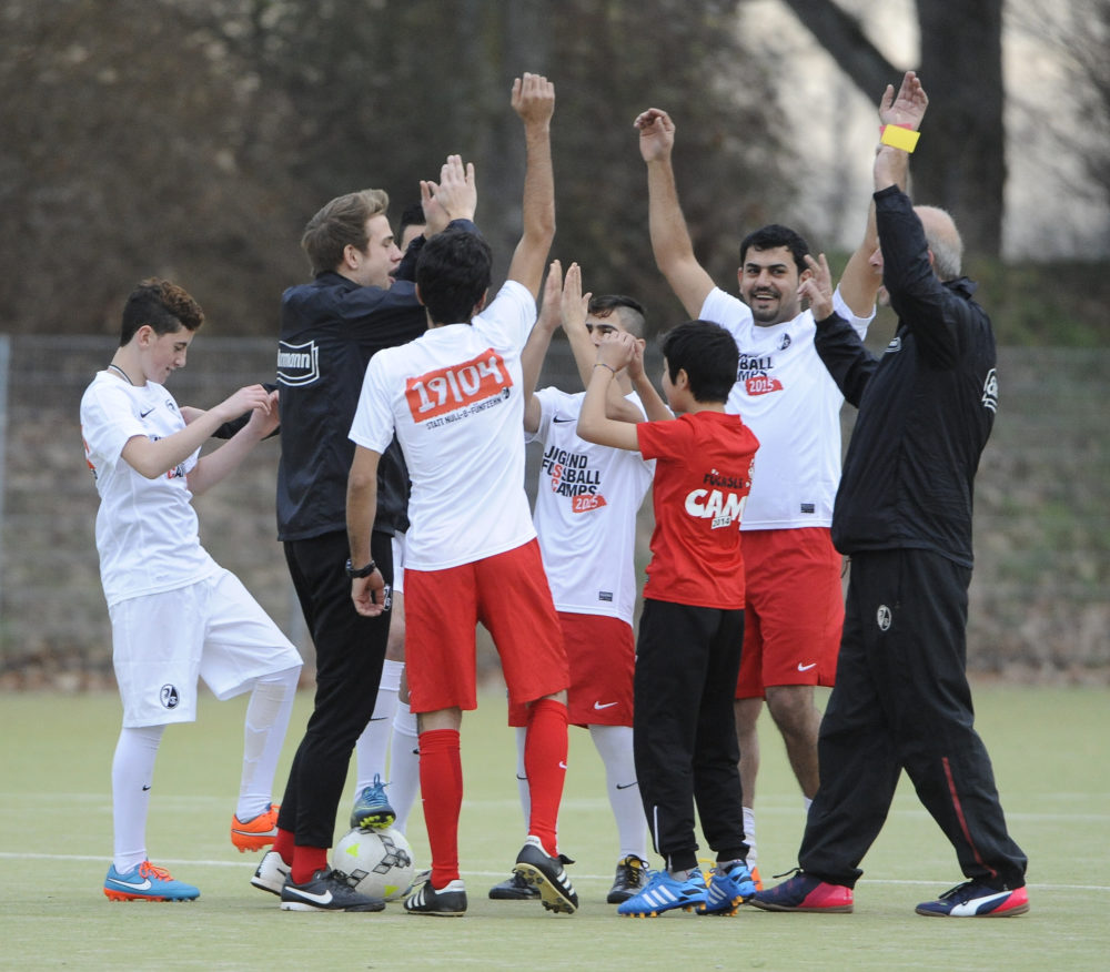Eine Gruppe von jungen Fußballspielern und ein Trainer stehen auf einem Fußballplatz und jubeln mit hoch erhobenen Händen. Die Spieler tragen weiße Trikots und Shorts, einige haben blaue oder rote Stutzen. Der Trainer trägt eine schwarze Jacke und Hose. Im Hintergrund ist grüne Vegetation zu sehen.