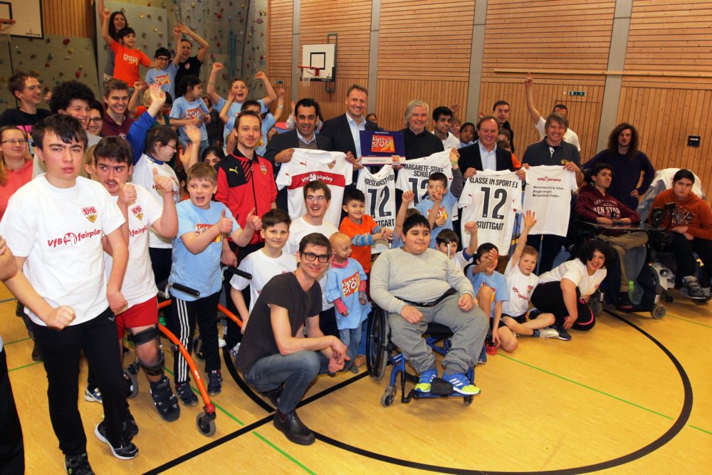 Eine Gruppe von Kindern und Jugendlichen, viele mit T-Shirts des VfB Stuttgart, posiert mit Betreuern und einem Mann im Anzug in einer Sporthalle. Einige der Kinder halten Schilder mit der Aufschrift "Klasse in Sport" und der Zahl "12".