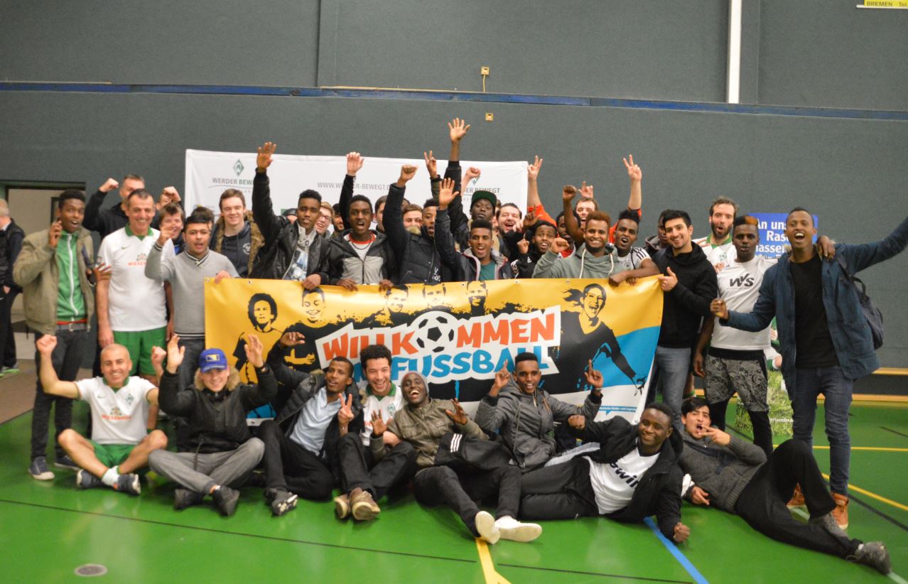 Eine große Gruppe von Menschen steht in einer Sporthalle zusammen und lächelt in die Kamera. Sie halten ein Banner mit der Aufschrift "WILLKOMMEN FUSSBALL" hoch. Einige der Personen haben Fußballtrikots an.