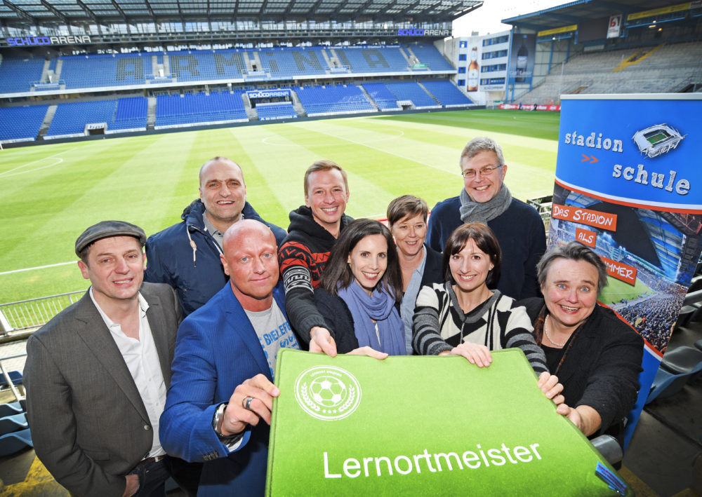 Eine Gruppe von etwa zehn Personen steht vor den Sitztribünen eines Fußballstadions. Sie halten ein grünes Schild mit der Aufschrift "Lernort Stadion" in den Händen. Im Hintergrund ist das Spielfeld und die Alm-Arena zu sehen.