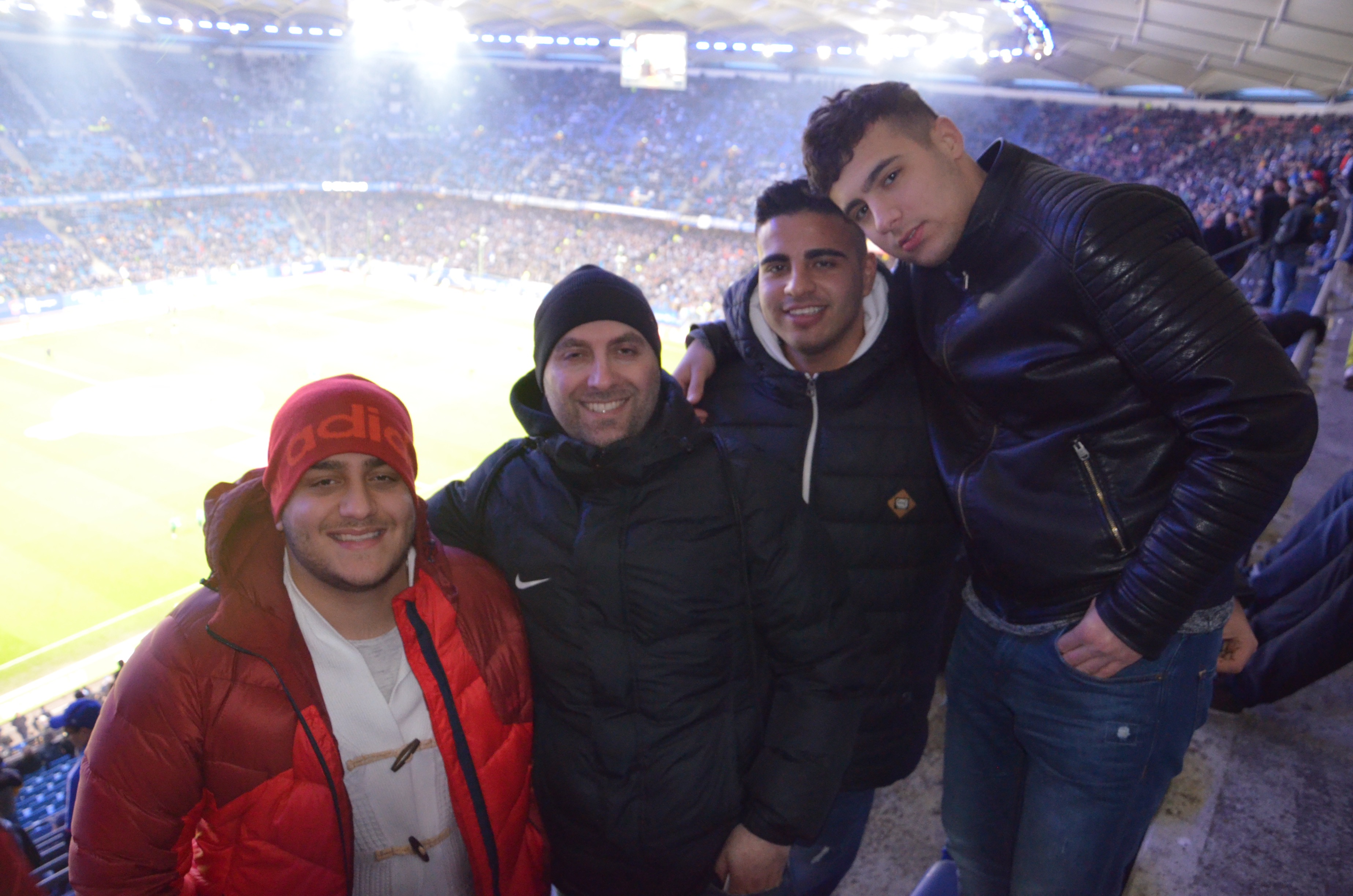 Vier junge Männer stehen lächelnd vor der Zuschauertribüne eines Fußballstadions. Im Hintergrund ist das beleuchtete Spielfeld zu sehen.