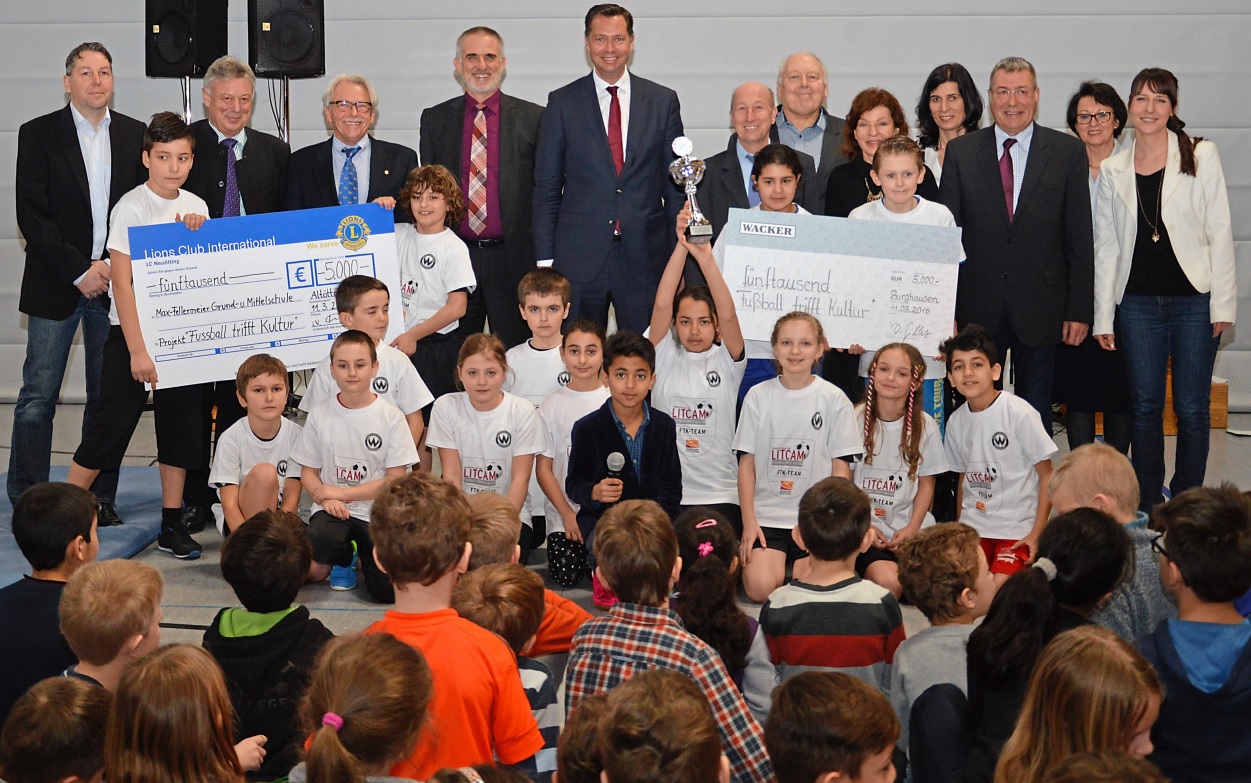 Eine Gruppe von Kindern in weißen T-Shirts mit dem Aufdruck "LITAN TEAM" posiert mit einem Pokal und Schecks vor einer Gruppe von Erwachsenen in einem Turnsaal. Die Schecks, überreicht vom Lions Club International, symbolisieren eine Spende für das Projekt "Fußball trifft Kultur". Im Hintergrund sitzen Zuschauer auf einer Tribüne.