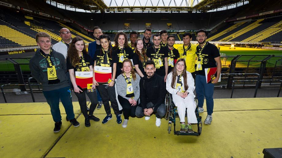 Eine Gruppe junger Menschen, viele mit gelben BVB-Schals, posiert auf einer Empore eines Fußballstadions. Im Hintergrund sind die gelben Sitze und der grüne Rasen des Spielfelds zu sehen. Eine Person sitzt im Rollstuhl.