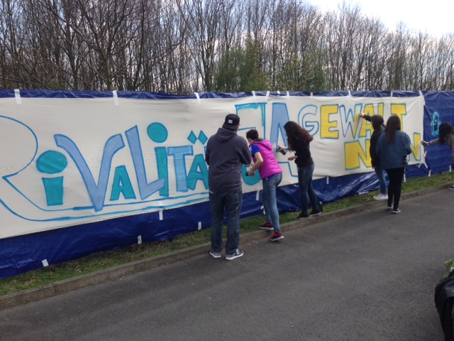 Eine Gruppe von Menschen malt ein Banner mit der Aufschrift "Rivalität – Ja, Gewalt – Nein". Im Hintergrund sind Bäume und eine Absperrung zu sehen.
