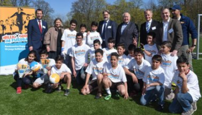 Eine Gruppe Jugendlicher steht auf einer Grünfläche vor einer Werbetafel mit dem Schriftzug "Fußball im Herzen". Hinter den Jugendlichen stehen erwachsene Männer und eine Frau, vermutlich Betreuer oder Vertreter der DFL Stiftung. Die Jugendlichen tragen weiße T-Shirts mit dem Aufdruck "Fußball im Herzen".