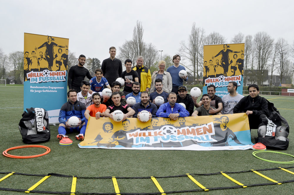 Eine große Gruppe von Menschen steht auf einem Fußballfeld und hält Fußbälle. Im Hintergrund sind zwei Banner mit der Aufschrift "Willkommen im Fußball - Bundesweites Engagement für junge Geflüchtete" zu sehen. Die Gruppe besteht aus Menschen unterschiedlichen Alters und Hintergrunds, darunter Fußballspieler und ehrenamtliche Helfer.