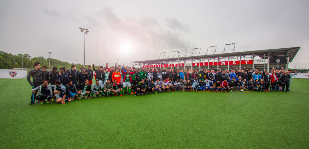Eine große Gruppe von Menschen steht auf einem grünen Fußballfeld und lächelt in die Kamera. Im Hintergrund ist ein Fußballstadion mit der Aufschrift "HEIMAT" zu sehen.
