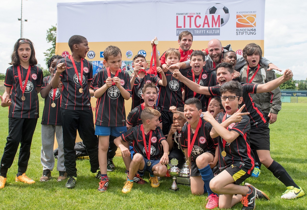 Jubelnde Kinder-Fußballmannschaft mit Medaillen und Pokal auf dem Rasen, im Hintergrund ein Banner mit dem Schriftzug "Fussball trifft Kultur" und dem Logo der Bundesliga-Stiftung.
