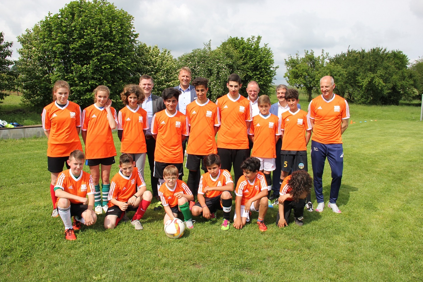 Eine Gruppe von Kindern und Jugendlichen in orange-weißen Fußballtrikots steht auf einer grünen Wiese und lächelt in die Kamera. Einige Erwachsene stehen hinter den Kindern. Im Vordergrund liegt ein Fußball.
