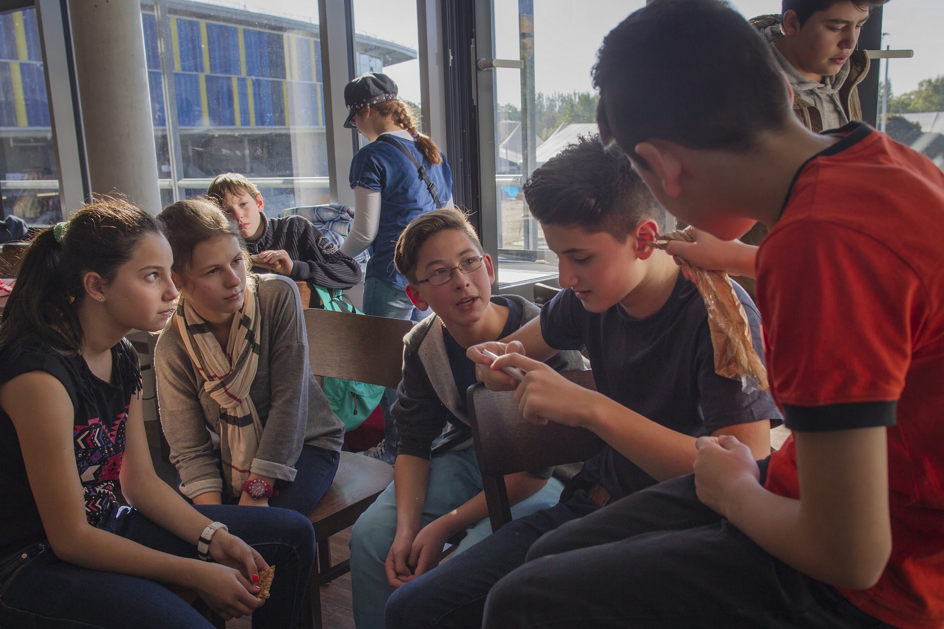 Eine Gruppe von Schülerinnen und Schülern sitzt in einer modernen, hellen Umgebung, vermutlich in einem Lernzentrum eines Fußballstadions, und macht Mittagspause. Einige unterhalten sich und zeigen auf ihre Handflächen, während andere im Hintergrund stehen und sich unterhalten.