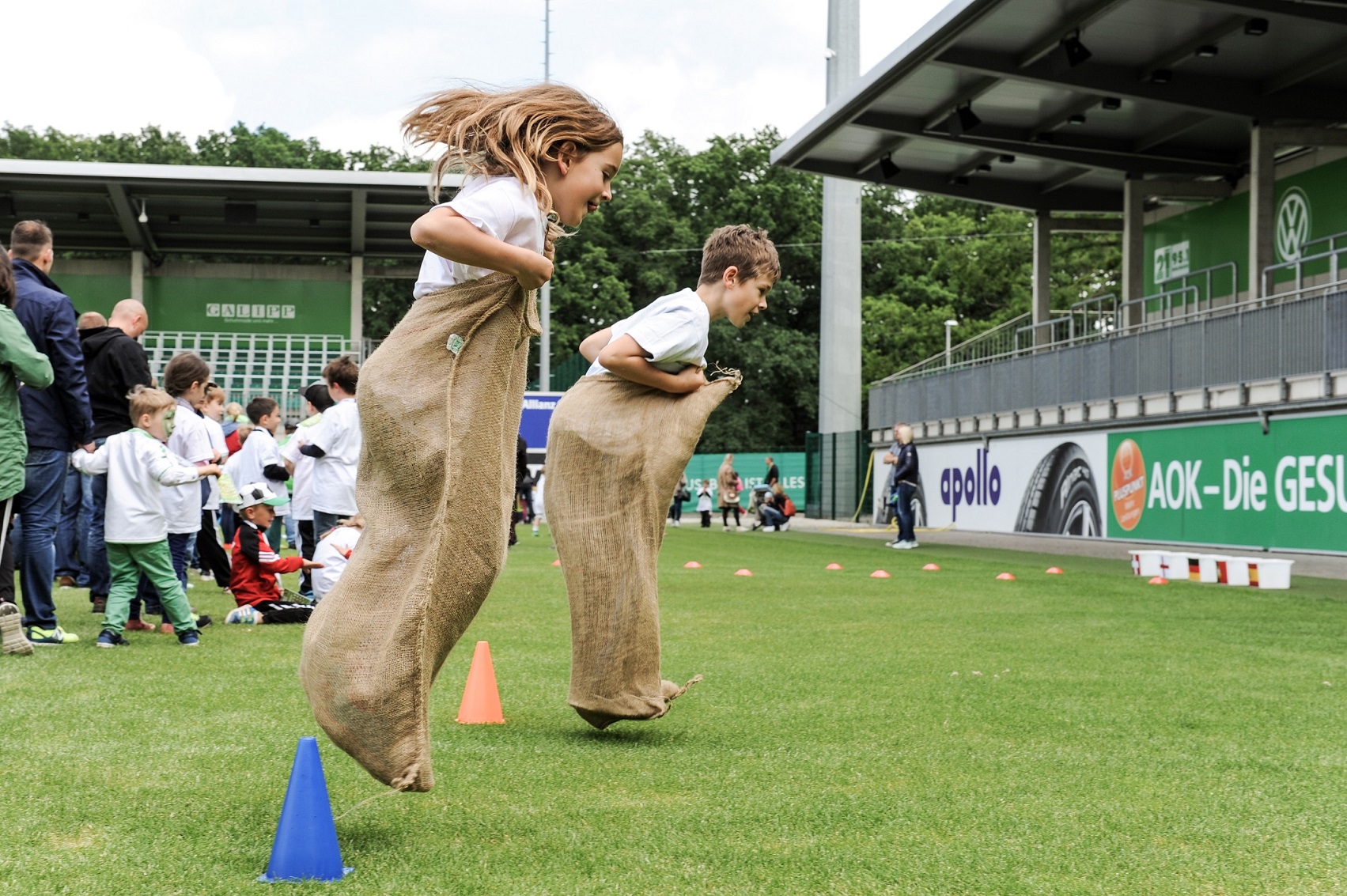 Zwei Kinder springen bei einem Sackhüpfen auf einem Fußballfeld, im Hintergrund eine Stadiontribüne und weitere Kinder beim Spielen.